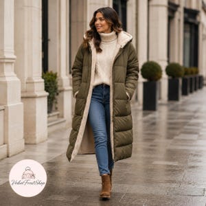 Può includere: Un lungo piumino verde oliva con cappuccio e fodera color crema. Il cappotto è indossato sopra un maglione color crema e jeans blu. La persona indossa stivali marroni. Il cappotto è lungo fino al ginocchio.