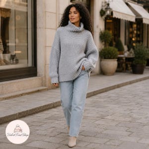 Puede incluir: Una mujer con un jersey de cuello alto de punto grueso gris claro y vaqueros azul claro. El jersey tiene un cuello alto y doblado y mangas largas. Lleva botines beige. El fondo muestra una calle de la ciudad con tiendas y una acera adoquinada.