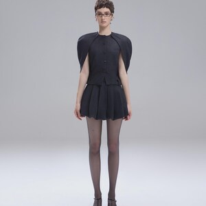 May include: A dark charcoal gray ensemble consisting of a button-front top with structured, cape-like sleeves and a matching pleated mini skirt. The outfit is paired with sheer black tights and black pointed-toe T-strap heels with thin ankle straps.
