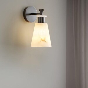Puede incluir: Aplique de pared moderno con acabado met&aacute;lico oscuro y pantalla blanca en forma de cono. La pantalla tiene un aspecto veteado y la luz es c&aacute;lida. El aplique est&aacute; montado en una pared gris.