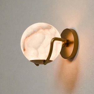 Puede incluir: Aplique de pared moderno con una pantalla esférica de alabastro blanquecino, con vetas cálidas de color marrón rojizo. El globo está sujeto por un brazo de metal curvado, con tono latón, y montado sobre una placa trasera circular. La lámpara proporciona una iluminación suave y ambiental.