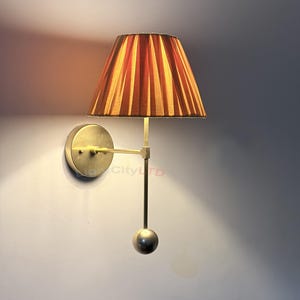 May include: A wall-mounted lamp featuring a pleated orange lampshade. The lamp has a brass finish, with a circular base and a straight arm extending to the shade. A small sphere hangs below the shade. The lamp is mounted on a light grey wall.