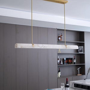 Op de afbeelding: Een moderne, rechthoekige hanglamp. De lamp heeft een marmerachtige, gebroken witte kap, opgehangen aan goudkleurige staven en een bijpassende plafondbevestiging. De lamp hangt boven een marmeren aanrecht.