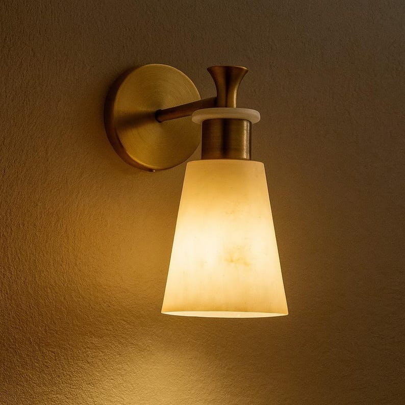 Puede incluir: Aplique de pared con acabado de lat&oacute;n y una pantalla c&oacute;nica de color blanquecino. El aplique tiene una placa trasera circular y un brazo curvo. La pantalla emite un brillo c&aacute;lido, ideal para la iluminaci&oacute;n ambiental.
