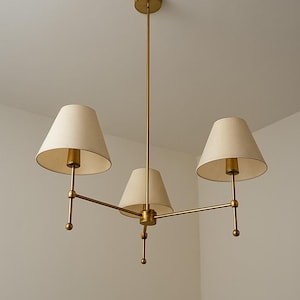 May include: A brass chandelier featuring three cream-coloured lampshades. The light fitting has a central vertical rod and horizontal arms extending to hold the shades. The shades are cone-shaped and the fixture is mounted on a white ceiling.