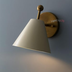 May include: A modern wall sconce with a conical off-white shade and a brass-colored base and arm. The sconce is mounted on a round brass plate. The light shines downward, creating a warm glow.