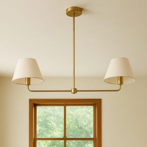 May include: A gold-toned, two-light pendant light fixture with off-white fabric shades. The fixture is suspended from the ceiling and has a horizontal bar with two downward-facing shades. The fixture is mounted above a wooden window.
