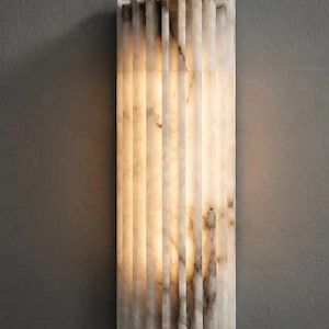 May include: A rectangular wall sconce featuring a vertical fluted design made of translucent white stone with natural grey veining. The fixture emits a soft, warm glow from within, highlighting the textured surface and the organic patterns of the stone.
