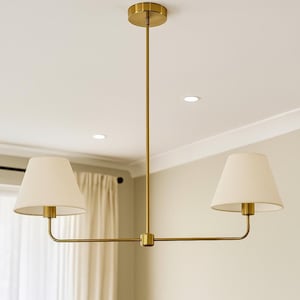 Puede incluir: Una lámpara de araña de dos luces con estructura de metal en acabado de latón cepillado, que presenta una varilla vertical central y dos brazos horizontales. Cada brazo sostiene una pantalla de tela color crema, creando un diseño equilibrado.