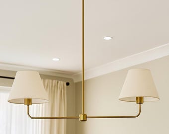 May include: A two-light chandelier featuring a brushed brass finish metal frame with a central vertical rod and two horizontal arms. Each arm supports a cream-colored, tapered fabric lampshade, creating a balanced and symmetrical lighting fixture.