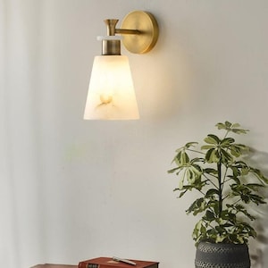 Puede incluir: Aplique de pared con acabado de lat&oacute;n y una pantalla blanca en forma de cono. El aplique est&aacute; montado en una pared clara. Una peque&ntilde;a planta en maceta y libros est&aacute;n sobre una superficie de madera debajo.