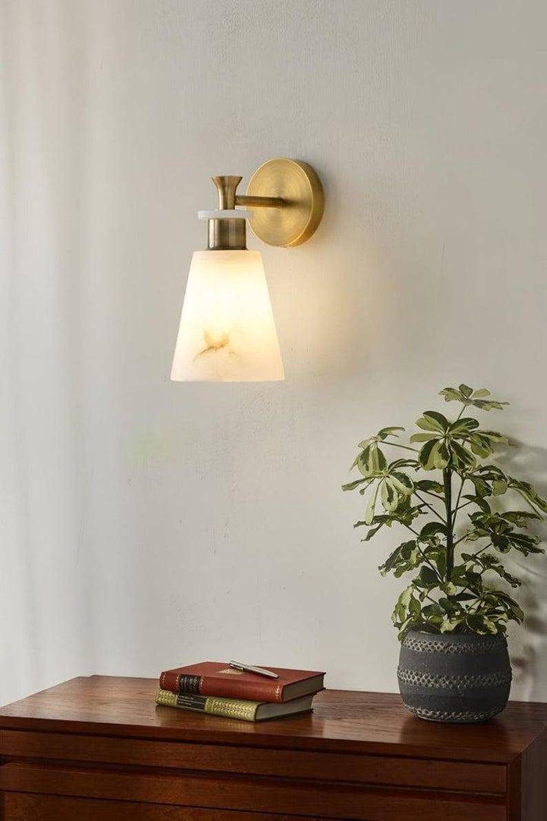 Puede incluir: Aplique de pared con acabado de lat&oacute;n y una pantalla blanca en forma de cono. El aplique est&aacute; montado en una pared clara. Una peque&ntilde;a planta en maceta y libros est&aacute;n sobre una superficie de madera debajo.