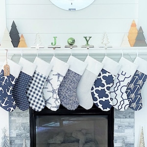 May include: A collection of nine Christmas stockings with various patterns, including anchors, polka dots, gingham, and geometric designs. The stockings are navy blue and white, hanging above a fireplace. Decorative wooden trees and the word "JOY" are on the mantel.