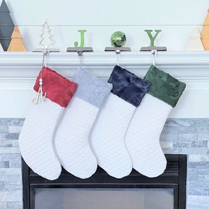 May include: Four white quilted Christmas stockings with plush cuffs in red, light blue, navy, and green hang above a fireplace. Decorative wooden trees and the word "JOY" are on the mantel. Each stocking has a silver hook.