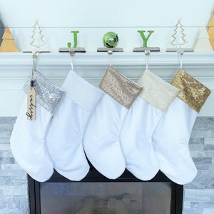 May include: Five white Christmas stockings hang above a fireplace, each with a different sequined cuff. The word "JOY" is spelled out above the stockings with green letters and a green bell. Decorative tree and wooden accents complete the festive scene.