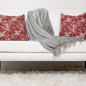 May include: A white couch with two red and white patterned throw pillows and a gray throw blanket with fringe. The couch is sitting on a white fluffy rug.