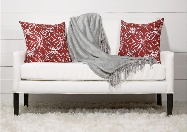 May include: A white couch with two red and white patterned throw pillows and a gray throw blanket with fringe. The couch is sitting on a white fluffy rug.