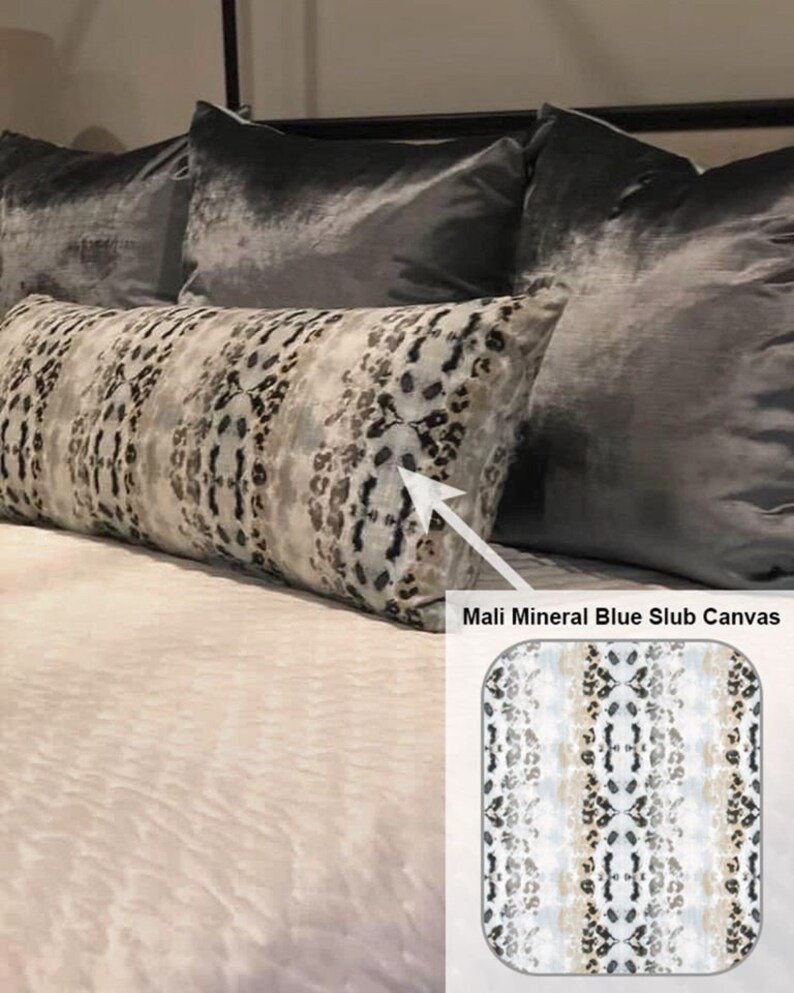 May include: A long, rectangular throw pillow with a white, gray, and black abstract animal print pattern. The pillow is on a bed with gray velvet pillows. The text "Mali Mineral Blue Slub Canvas" is visible below the pillow.