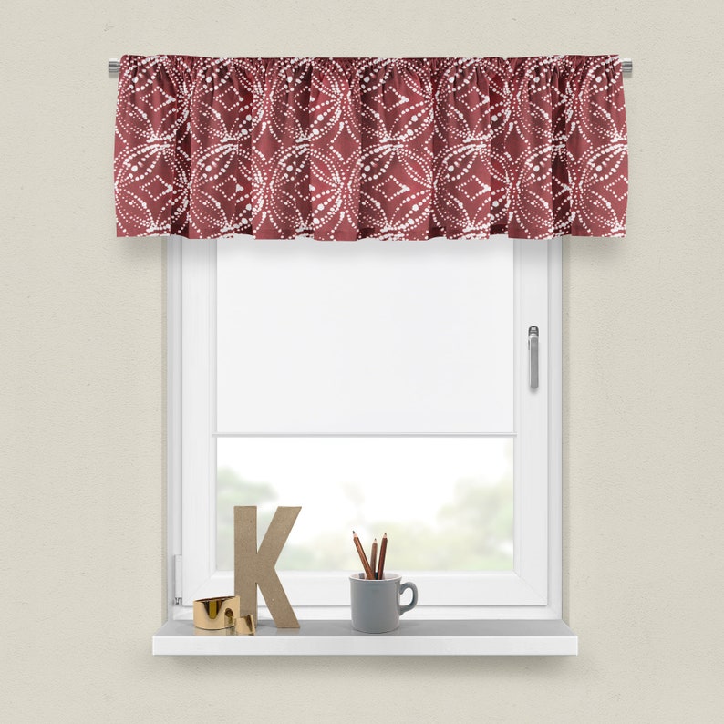 May include: A red valance with a white geometric pattern. The valance is hung on a white window with a white window sill. There is a wooden letter K, pencils in a gray cup, and a gold ring on the window sill.