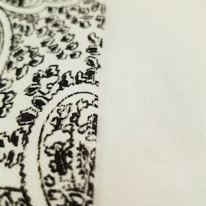 Op de afbeelding: Close-up van een witte stof met een zwart paisley-patroon. De stof is glad en heeft een subtiele glans.