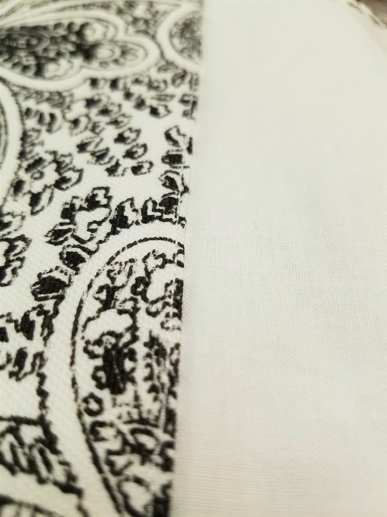 Op de afbeelding: Close-up van een witte stof met een zwart paisley-patroon. De stof is glad en heeft een subtiele glans.