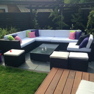 Op de afbeelding: Een zwarte wicker patio-hoekbank met witte kussens en kleurrijke kussens. De bank is in een L-vorm gerangschikt en bevat een salontafel en verschillende poefs.