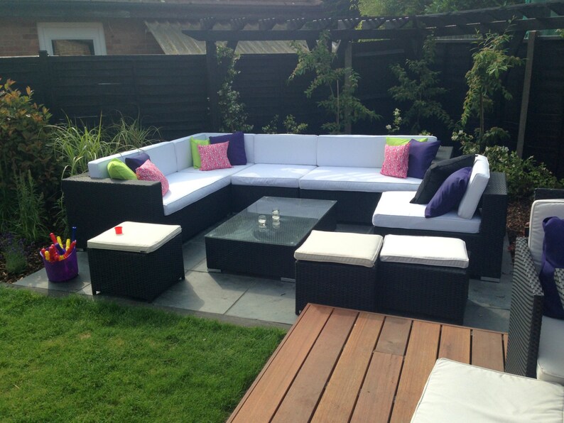 Op de afbeelding: Een zwarte wicker patio-hoekbank met witte kussens en kleurrijke kussens. De bank is in een L-vorm gerangschikt en bevat een salontafel en verschillende poefs.