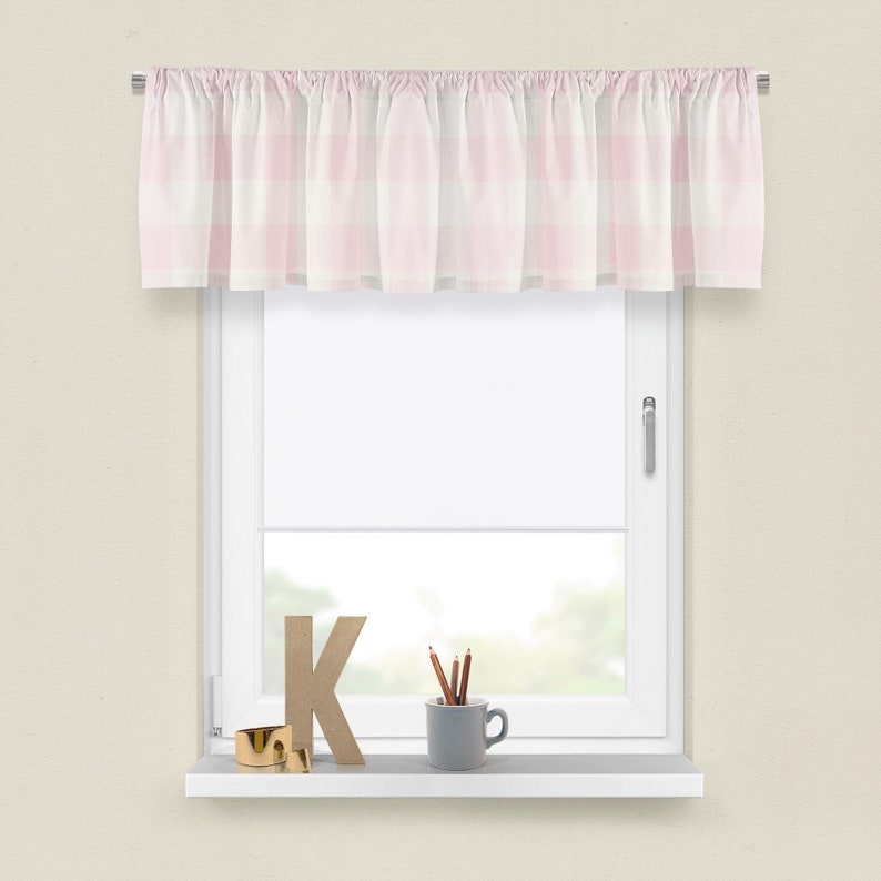 May include: A pink and white gingham valance with frills hangs on a window. The window is white and has a white sill. On the sill are a wooden letter K, a cup with pencils, and a gold ring.