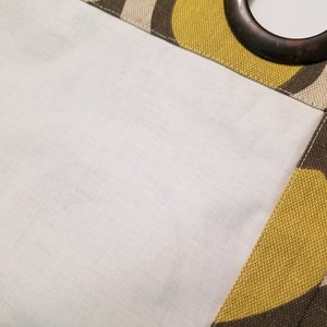 Op de afbeelding: Close-up van een bruin, wit en geel gepatroneerd gordijn met een bruine metalen oog. Het gordijn is gevoerd met een witte stof.