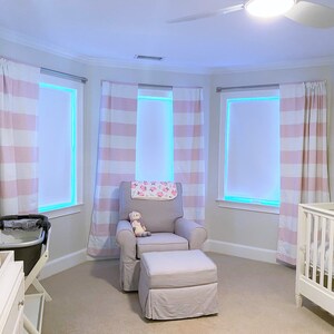 May include: A nursery room with a white cot, a rocking chair with a pink and white striped throw, and a white chest of drawers with a changing mat. The room has three windows with pink and white striped curtains.