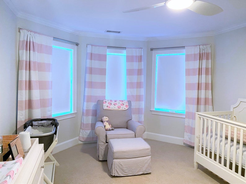 May include: A nursery room with a white cot, a rocking chair with a pink and white striped throw, and a white chest of drawers with a changing mat. The room has three windows with pink and white striped curtains.