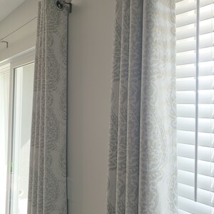 May include: Two white curtains with a grey patterned design hang from a silver curtain rod. The curtains are drawn closed, and the pattern is visible on the fabric.