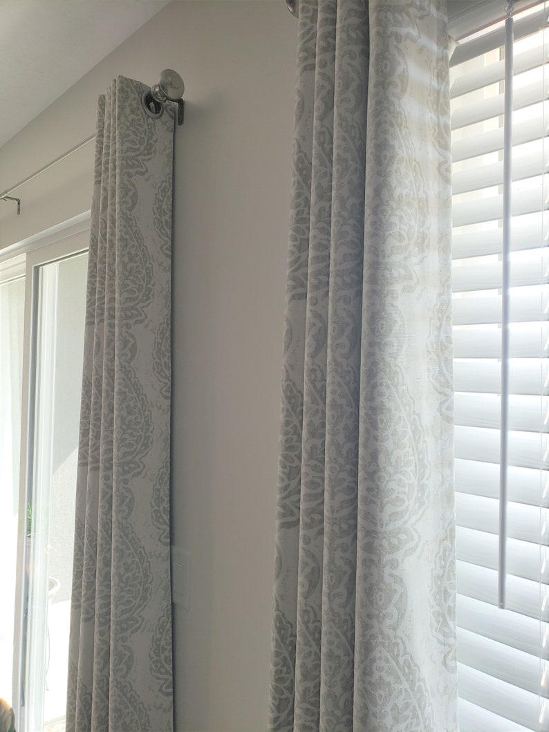 May include: Two white curtains with a grey patterned design hang from a silver curtain rod. The curtains are drawn closed, and the pattern is visible on the fabric.