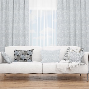 May include: A white sofa with a throw blanket and several patterned pillows in a living room with light gray curtains and a window with a view of a blue sky.