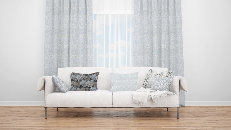 May include: A white sofa with a throw blanket and several patterned pillows in a living room with light gray curtains and a window with a view of a blue sky.