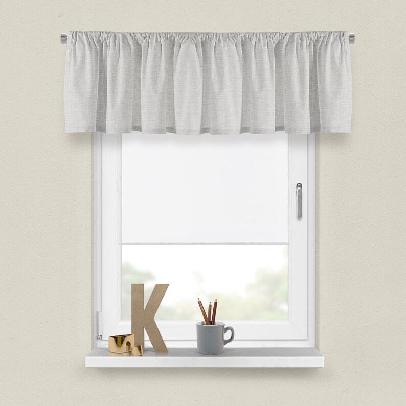 May include: A white valance with a ruffled edge hangs above a window with a white window frame. The windowsill is white and has a wooden letter K, a small gold ring, and a grey ceramic mug with pencils inside.