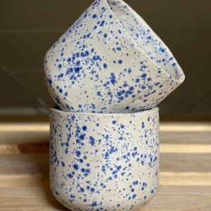 Pode incluir: Duas canecas de cerâmica empilhadas. As canecas são brancas com padrões salpicados em azul. A caneca inferior é cilíndrica, enquanto a caneca superior tem um design angular único. As canecas estão colocadas numa superfície de madeira.