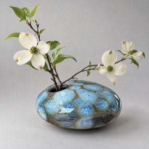 May include: A low, rounded ceramic vase with a glossy, crystalline glaze featuring blue and brown circular patterns. A single branch with white, four-petaled dogwood flowers and green leaves emerges from the small opening at the top.