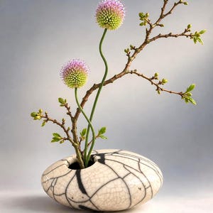 May include: A low, rounded ceramic vase with a cream-colored crackle glaze and bold black web-like patterns. Two spherical, light purple and green allium flowers on long, slender stems and a bare branch with small green leaves emerge from it.