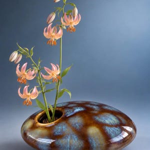 May include: A low, rounded ceramic vase with a glossy brown and blue mottled glaze. The vase features a single circular opening containing a metal flower frog holding a stem of tall, light orange lily flowers with green leaves.