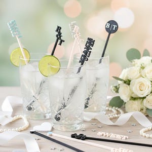 May include: Custom stir sticks in glasses with lime slices. The sticks are black, white, and light blue, with various designs including initials and names. The image also features a bouquet of white roses and decorative elements.