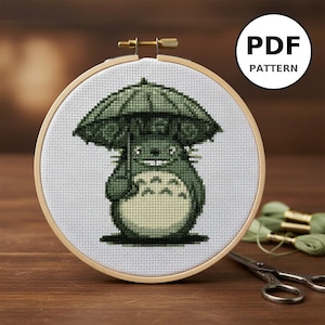 Może przedstawiać: Wzór haftu krzyżykowego przedstawiający zieloną postać Totoro z parasolem, umieszczony w drewnianej tamborku. Wzór znajduje się na białej tkaninie, z napisem "PDF PATTERN" w białym okręgu. Widoczne są nożyczki i zielona nić.