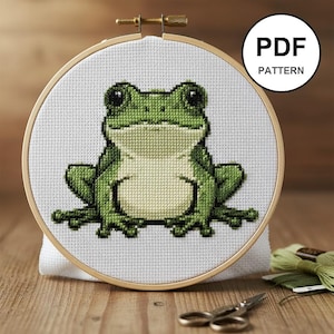 Peut inclure: Un motif de point de croix d'une grenouille verte dans un cercle à broder en bois. La grenouille est centrée sur un tissu blanc, avec le texte "PDF PATTERN" dans un cercle blanc. Ciseaux et fil vert visibles.