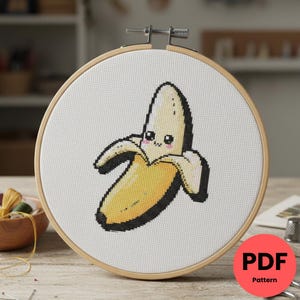 May include: Cross-stitch design of a smiling banana, with a yellow peel and black outline, set in a wooden embroidery hoop. The design is on white fabric. A red circle with the text "PDF Pattern" is in the lower right corner.