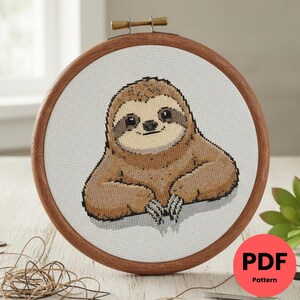 以下が含まれることがあります： 木製の刺繍枠に入った、笑顔のナマケモノのクロスステッチパターン。ナマケモノは茶色とタン色で、顔は薄いベージュ、目は濃い色です。「PDF Pattern」という文字が赤い円の中にあります。