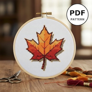 Peut inclure: Un motif de point de croix d'une feuille d'érable aux tons orange et rouge, présenté dans un cercle à broder en bois. Le texte "PDF PATTERN" est dans un cercle en haut à droite. Ciseaux et fil à broder sur une surface en bois.