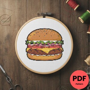 Peut inclure: Un motif de point de croix d'un hamburger pixélisé, présenté dans un cercle à broder en bois. Le burger comprend un pain aux graines de sésame, de la laitue, de la tomate, de l'oignon rouge, du fromage et un steak haché. Le motif PDF est visible.
