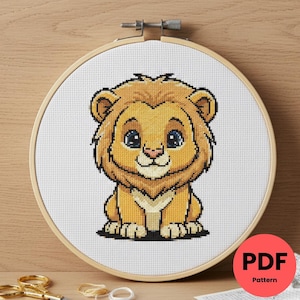 Peut inclure: Un motif de point de croix d'un lionceau de dessin animé dans un cercle à broder en bois. Le lionceau est jaune et beige avec des yeux bleus et un contour noir. Le motif est sur tissu blanc. Un cercle rouge avec le texte "PDF Pattern" est en bas à droite.