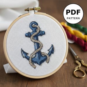 Peut inclure: Un motif de point de croix d'ancre bleue avec un détail de corde est présenté dans un cercle à broder en bois. Le texte "PDF PATTERN" est dans un cercle noir. Du fil à broder coloré et des ciseaux dorés sont en arrière-plan.
