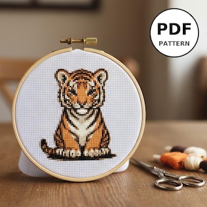 Peut inclure: Une broderie au point de croix d'un bébé tigre dans un cercle à broder. Le tigre est orange et noir, avec une poitrine et un ventre blancs. Le cercle est en bois. Le texte "PDF PATTERN" est dans un cercle en haut à droite.